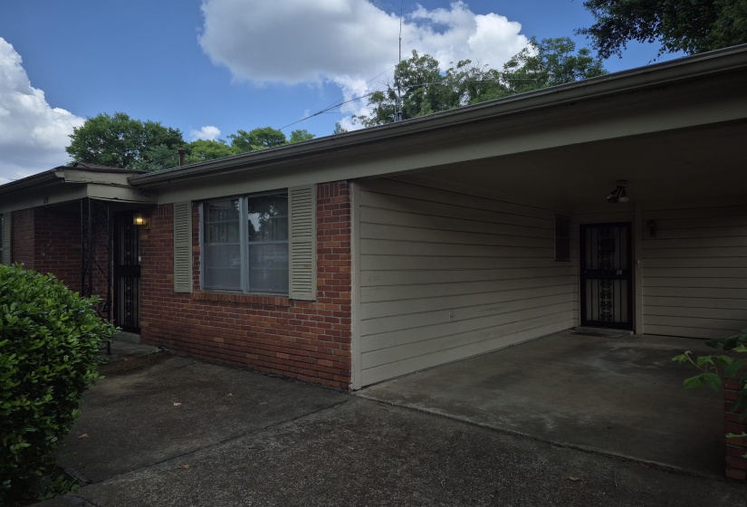 Brick exterior with an attached carport, featuring concrete paving, exterior lighting, and a security screen door