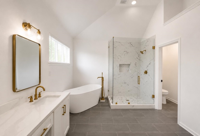 Spacious bathroom featuring a frameless glass shower with marble-look tile, a freestanding soaking tub, and a vanity with a white countertop and brass-finish fixtures