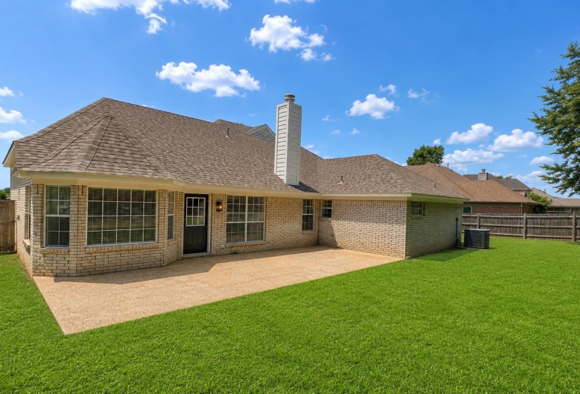 Expansive backyard featuring a large aggregate patio, a spacious green lawn, a brick exterior with multiple windows, and a prominent chimney structure
