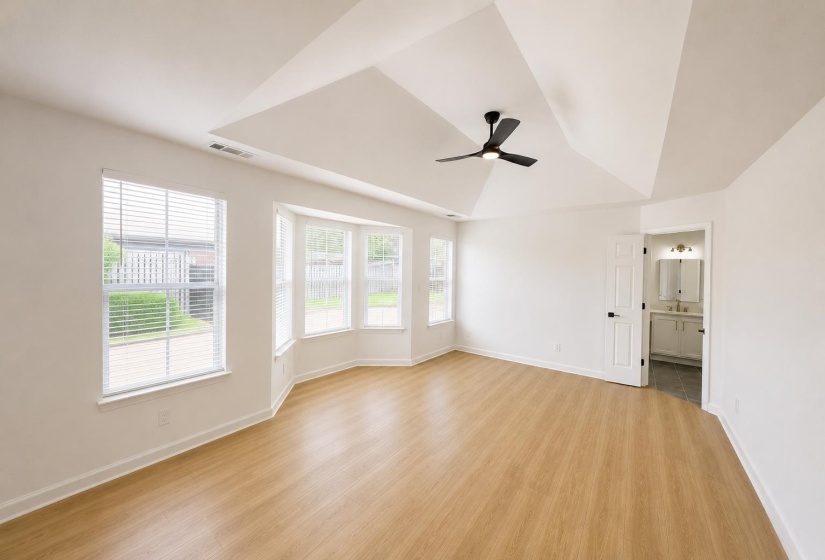 Spacious room featuring a tray ceiling with a black ceiling fan, wood-finish flooring, a bay window, and a door opening to a bathroom with a vanity and mirror