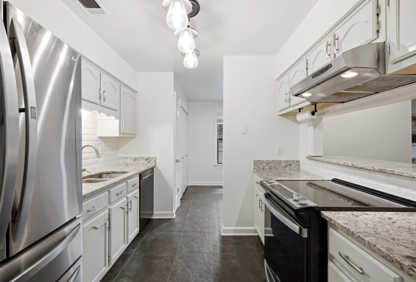 Galley kitchen featuring light-toned cabinetry, stainless steel appliances, granite countertops, a subway tile backsplash, and dark tile flooring