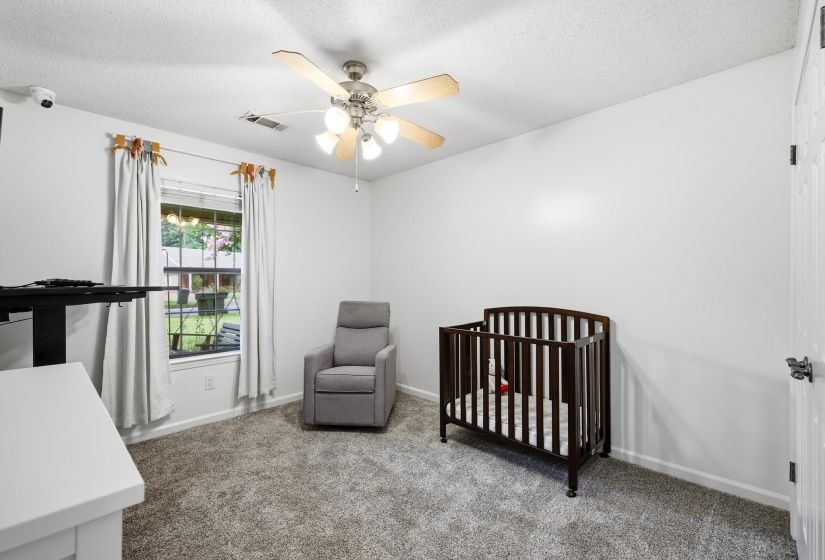 Bedroom featuring neutral wall tones, textured carpeting, a multi-pane window with exterior landscaping visible, a ceiling fan with integrated lighting, and a paneled door