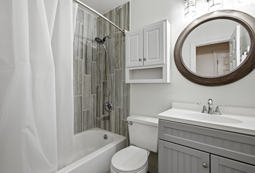 Full bathroom featuring a white bathtub, gray-toned rectangular tile shower surround, a gray vanity with beadboard detailing, a white countertop with integrated sink, and an oval mirror with a decorative frame