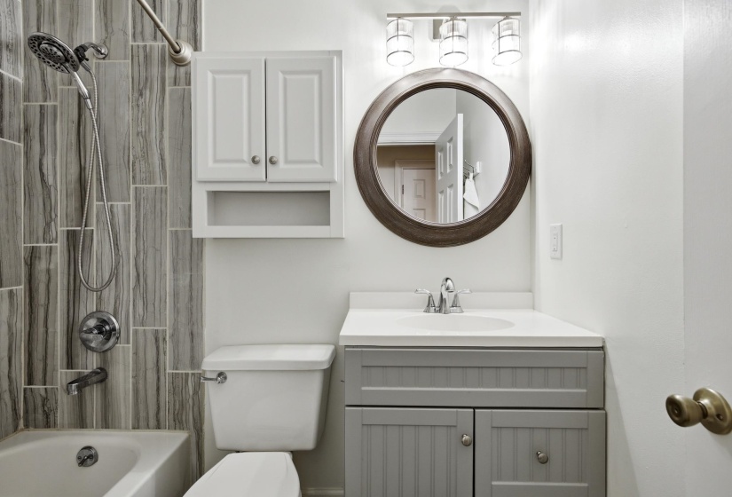 Bathroom featuring a tub/shower combination with vertically-stacked gray-toned plank tile surround, a white vanity with a light countertop, a round wood-framed mirror, and a three-light fixture