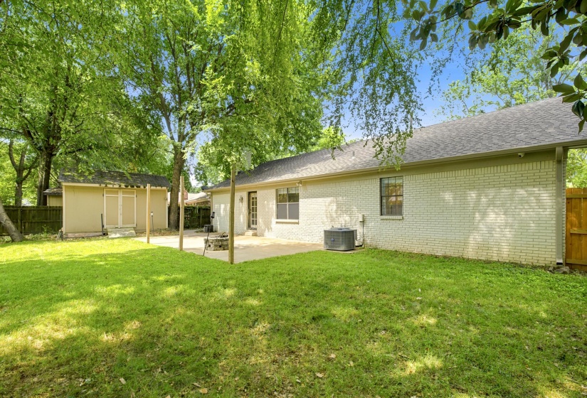 Spacious backyard featuring a large grass area, mature trees providing natural shade, a concrete patio, a detached storage shed, and a privacy fence