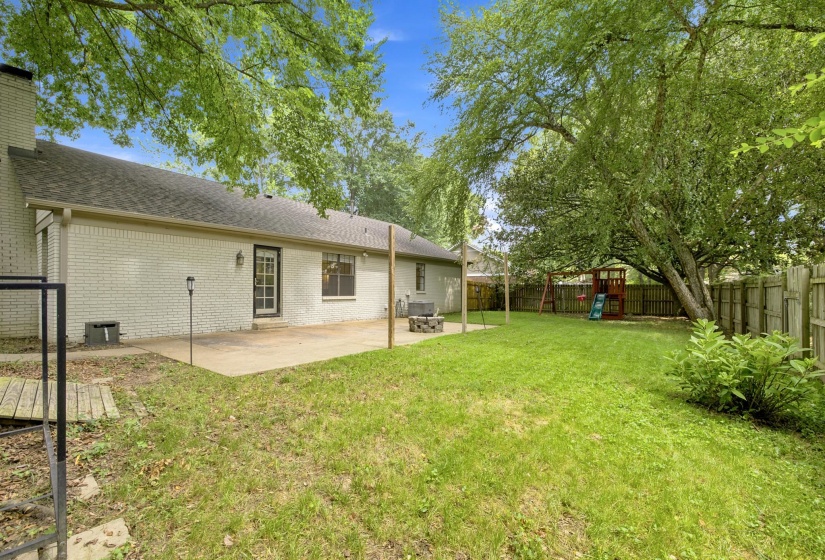 Expansive backyard with a large concrete patio, mature trees providing shade, and a wood privacy fence