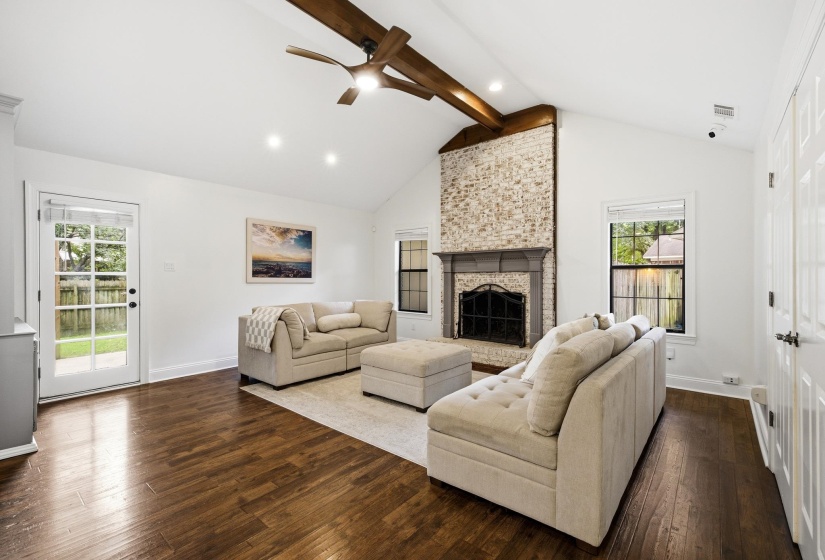 Vaulted ceiling living area featuring wood-finish flooring, a painted brick fireplace with a carved mantel, exposed wood beams, and a paneled glass door opening to the exterior