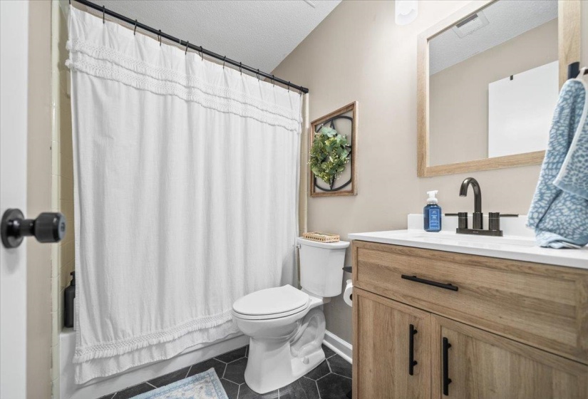 Bathroom featuring a wood-finish vanity with a white countertop, black hexagonal floor tiles, a framed mirror, and a built-in tub with a shower