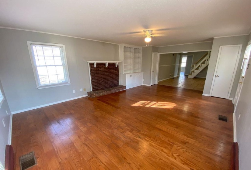 Spacious living area featuring polished wood-finish flooring, a brick fireplace with a white mantel, built-in shelving, and a ceiling fan