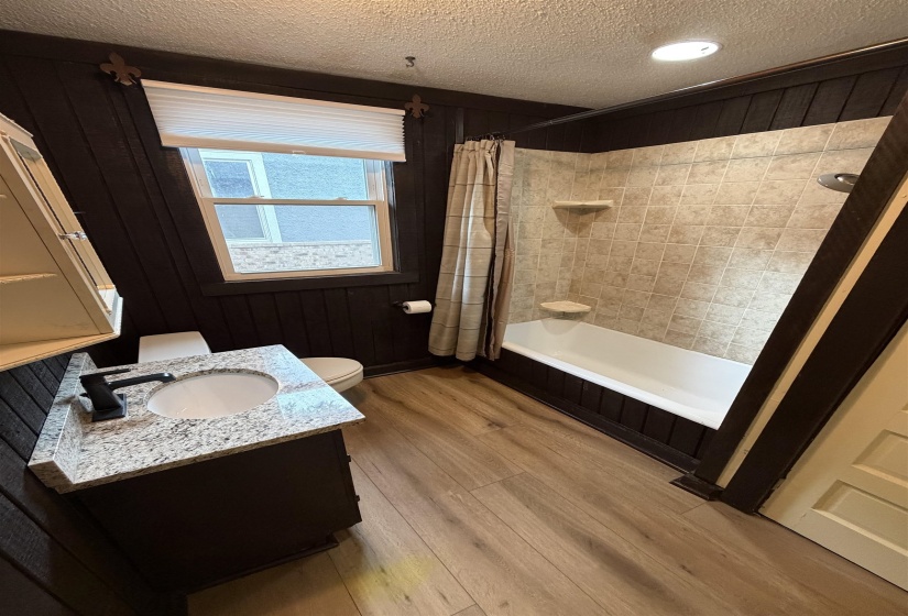 Full bathroom featuring a single window, a dark wood-finish vanity with a granite-style countertop, a rectangular undermount sink with a matte black faucet, a white toilet, a shower/tub combination with tiled surround, and wood-finish flooring