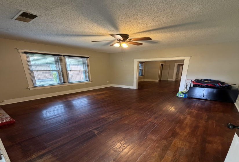 Spacious room featuring rich wood-finish flooring, a ceiling fan with light fixture, and natural light from a double window with blinds