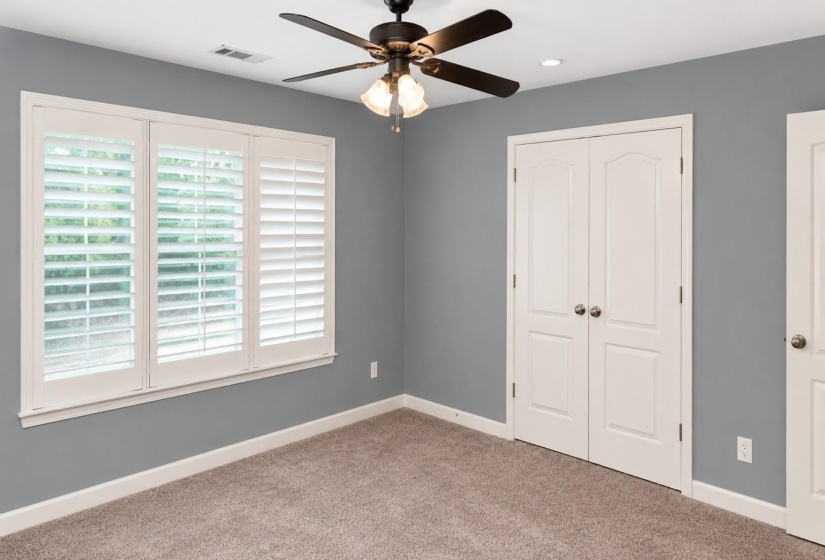 Carpeted room featuring a ceiling fan with light fixture, three-panel window with white plantation shutters, and double bi-fold doors