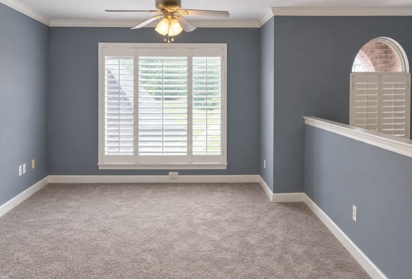 Spacious room featuring neutral carpeting, white plantation shutters, a ceiling fan with light fixture, and crown molding