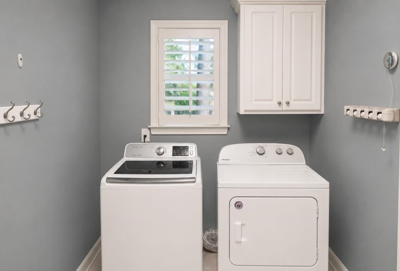 Laundry room featuring a white-framed window with plantation shutters, overhead white cabinetry, and light gray walls