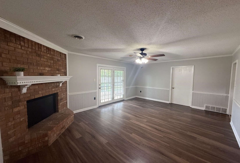 Spacious living area featuring a brick fireplace with a decorative mantel, wood-finish flooring, and light gray walls with wainscoting