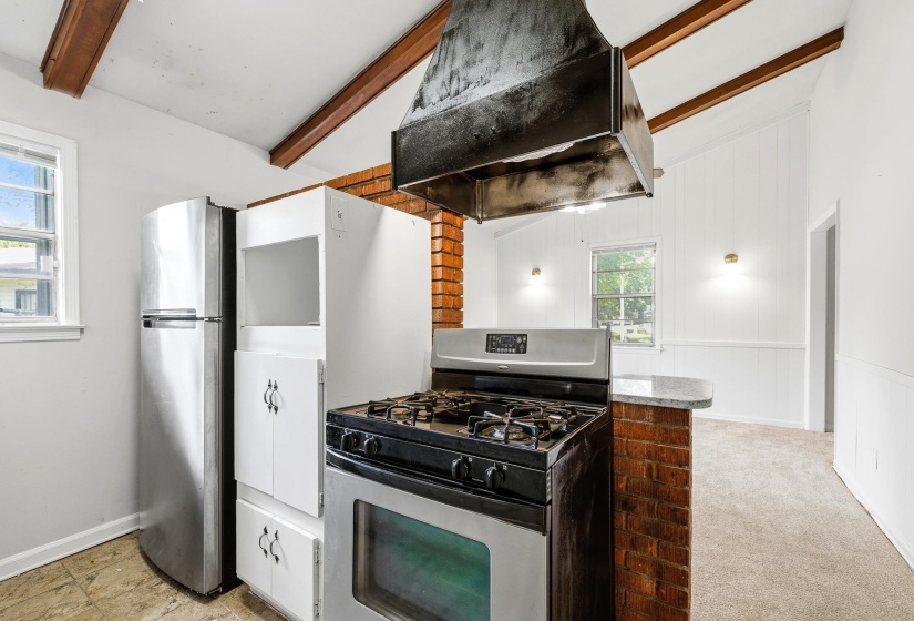 Kitchen featuring a stainless steel range, brick island with countertop seating, exposed wood ceiling beams, vaulted ceiling, and built-in white cabinetry