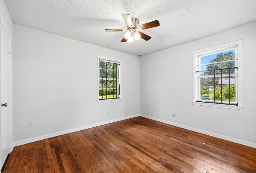 Room featuring two windows with security bars, a ceiling fan with integrated lighting, wood-finish flooring, and white painted walls
