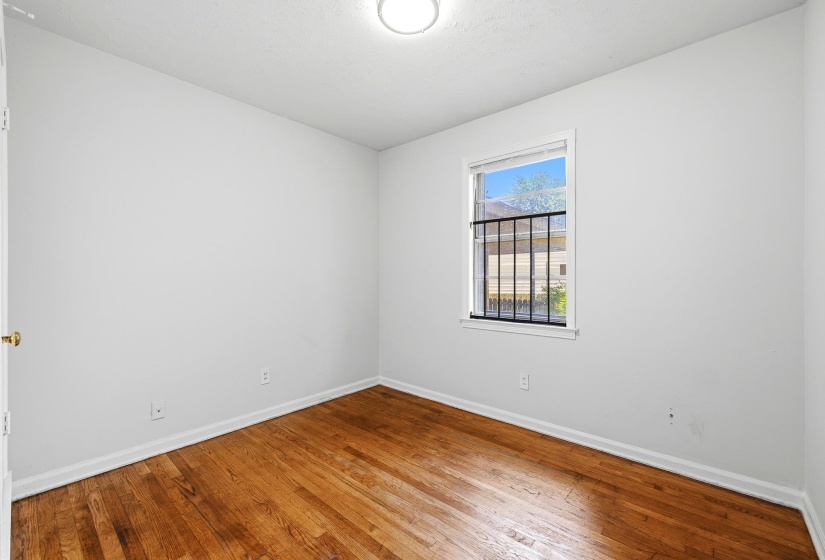 Hardwood flooring, white painted walls, white baseboards, and a single window with security bars