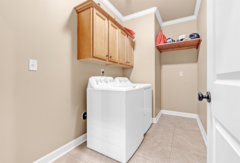 Dedicated laundry area featuring light wood-finish cabinetry, a built-in wood-finish shelf, and neutral-toned floor tiling