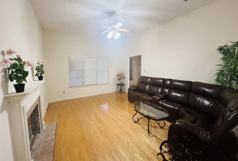 Living area featuring wood-finish flooring, a white fireplace with a brick hearth, a window with horizontal blinds, and a ceiling fan