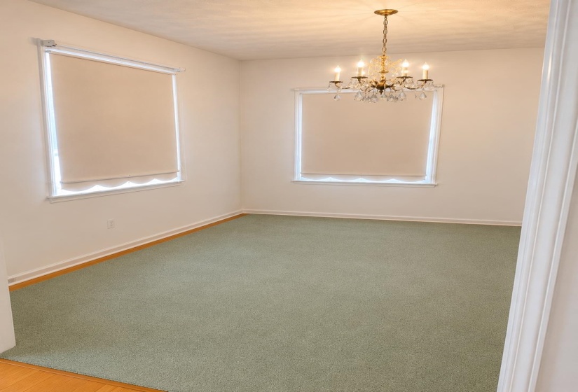 Room featuring light green carpeting, two windows with roller shades, and a brass-finish chandelier