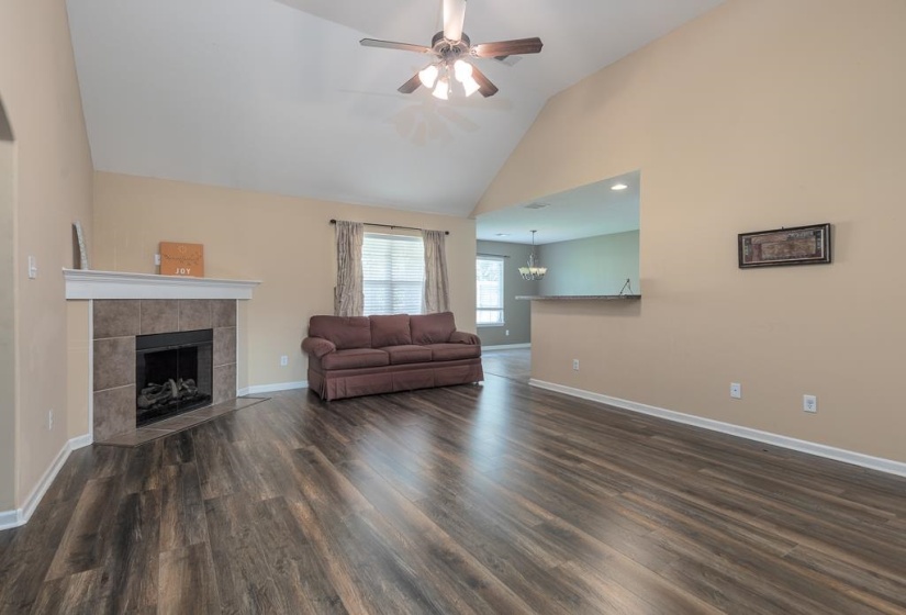 Living room featuring a vaulted ceiling with a ceiling fan, wood-finish flooring, a tiled fireplace with a mantel, and a pass-through to eat-in kitchen and another entrance to Bedrooms 2 and 3.