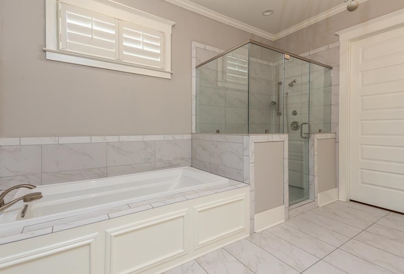 Soaking tub with white paneled surround, glass-enclosed shower, white tile flooring, and a window with plantation shutters
