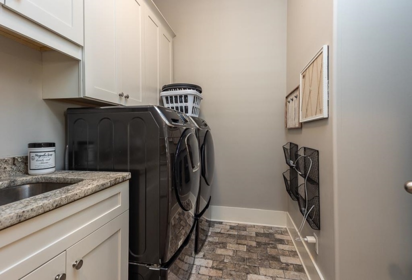 Utility room featuring a built-in sink with a granite-style countertop, white cabinetry with silver-tone hardware, and a stone-look floor