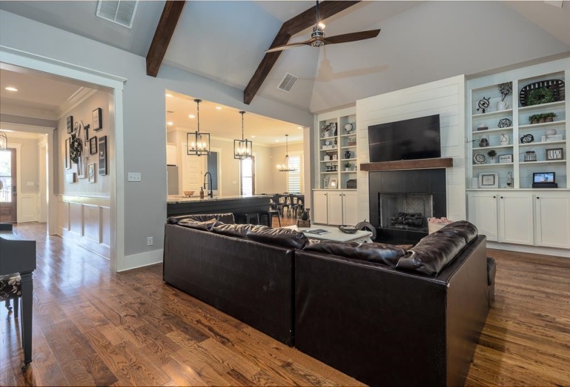 Spacious living area featuring high vaulted ceilings with exposed wood beams, wood-finish flooring, a fireplace with a dark surround and wood mantel, and built-in shelving with lower cabinetry