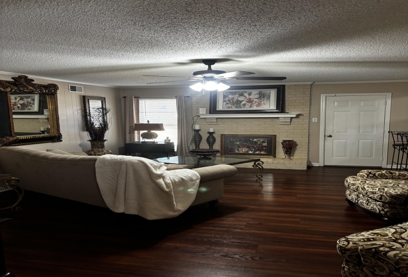 Living area featuring dark wood-finish flooring, a brick fireplace with mantel, and a ceiling fan with integrated lighting