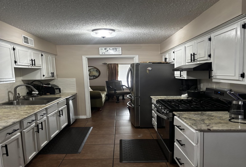 Galley kitchen featuring white cabinetry with matte black hardware, stone-finish countertops, stainless steel appliances, a tile backsplash, and tile flooring