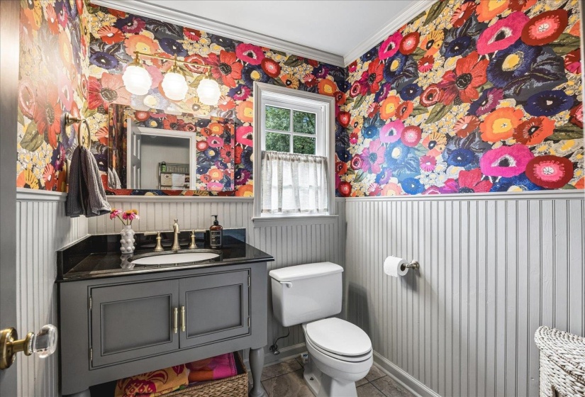 Powder room featuring vibrant floral wallpaper, beadboard wainscoting, a dark gray vanity with a stone countertop, a rectangular mirror, and a window with natural light