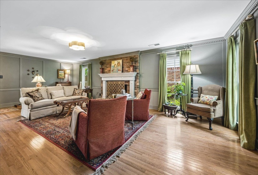 Living room featuring wood-finish flooring, a brick fireplace with a white mantel, paneled walls, crown molding, and multiple windows