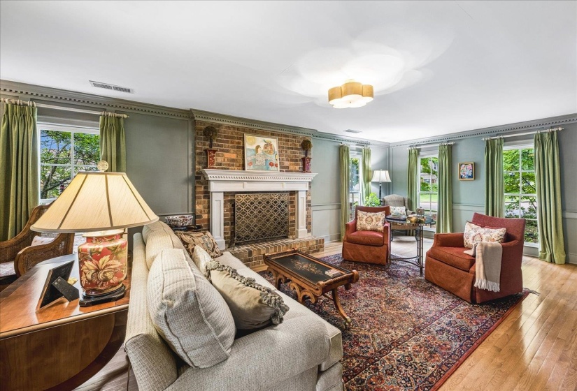 Spacious living room featuring a brick fireplace with a white mantel, hardwood flooring, multiple large windows, detailed crown molding, and a central ceiling light fixture