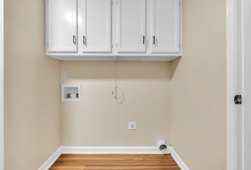 Dedicated utility space featuring overhead white cabinetry with dark pulls, light beige walls, and wood-finish flooring