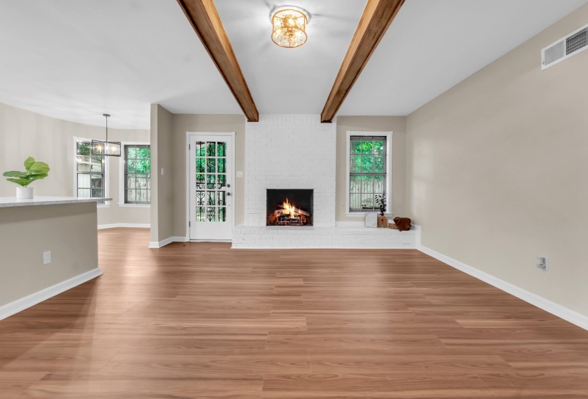 Spacious living area featuring wood-finish flooring, a white brick fireplace with a hearth, a glass-paned door, and two exposed ceiling beams
