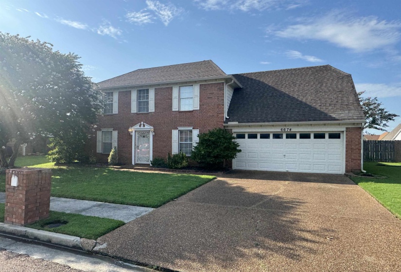 Brick facade residence featuring a two-car garage, white window shutters, and a mature front yard tree