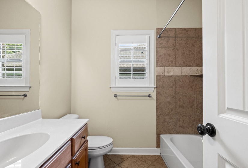 Bathroom featuring a vanity with a white countertop and integrated sink, a built-in tub with brown tile surround and accent band, a window with plantation shutters, and diamond-patterned floor tiling