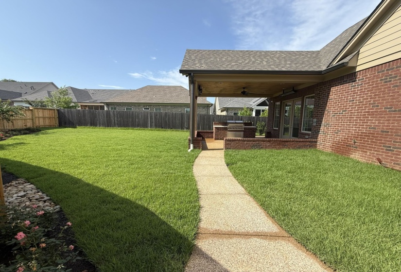 Expansive backyard featuring a covered patio, built-in outdoor kitchen, brick exterior, and a manicured lawn with a concrete pathway