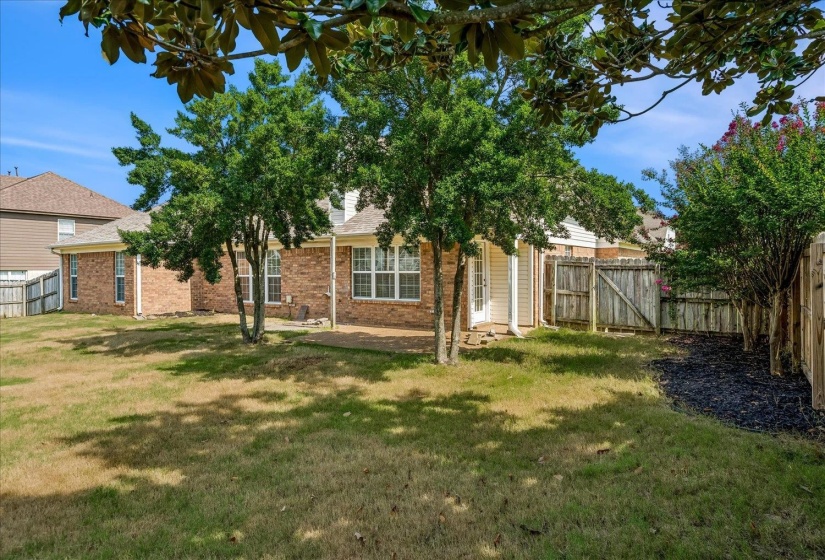 Expansive backyard featuring a partial privacy fence, mature trees, and a brick exterior with white-framed windows