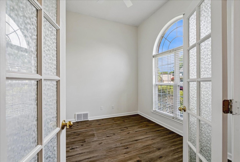 French doors with textured glass panels open to a room featuring an arched window, white blinds, and wood-finish flooring