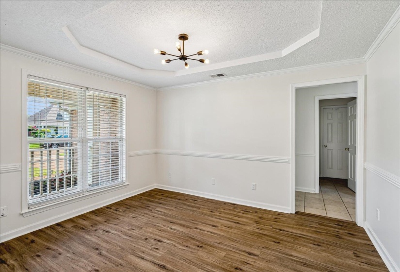 Spacious room featuring wood-finish flooring, a tray ceiling with a modern light fixture, and a large window with blinds
