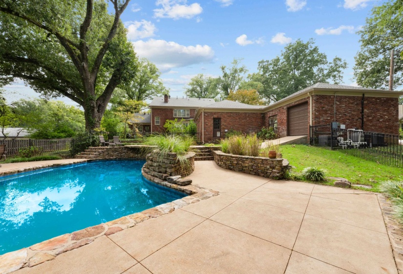 Fenced pool with expansive scored concrete deck, weeping stone wall, stone waterfall and stone planters.