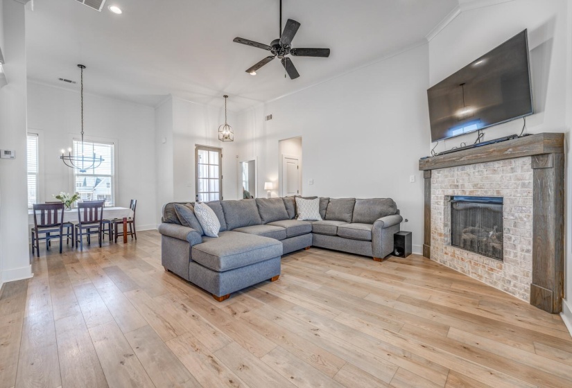 Living area featuring light wood-style flooring, a brick fireplace, baseboards, ornamental molding, and ceiling fan with notable chandelier