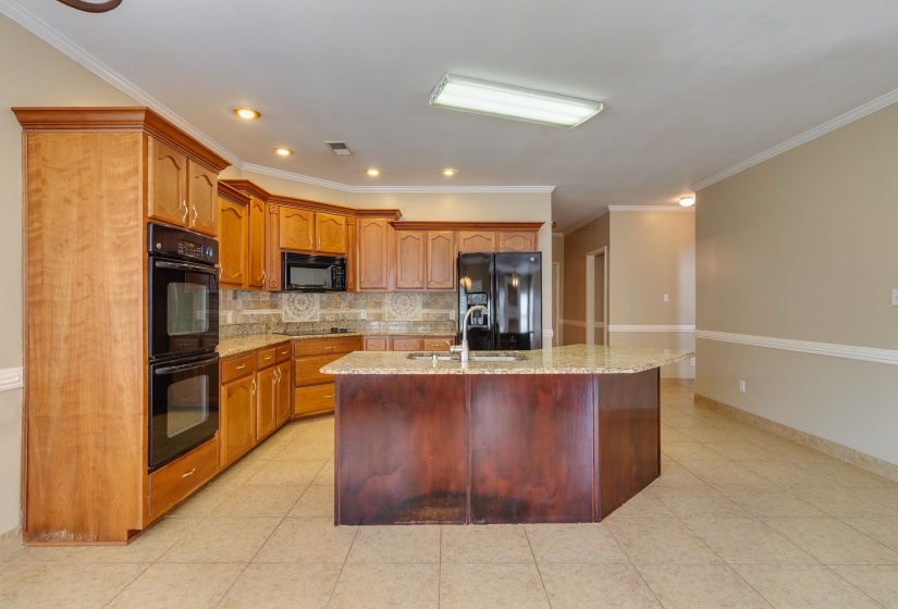 Kitchen with tasteful backsplash, ornamental molding, black appliances, light stone countertops, and a kitchen island with sink