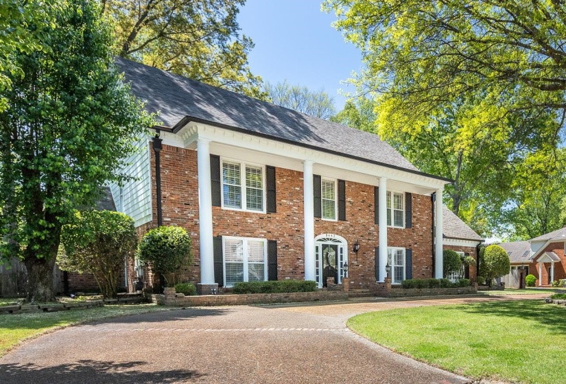 Neoclassical home with brick siding, covered porch, a front yard, driveway, and a shingled roof