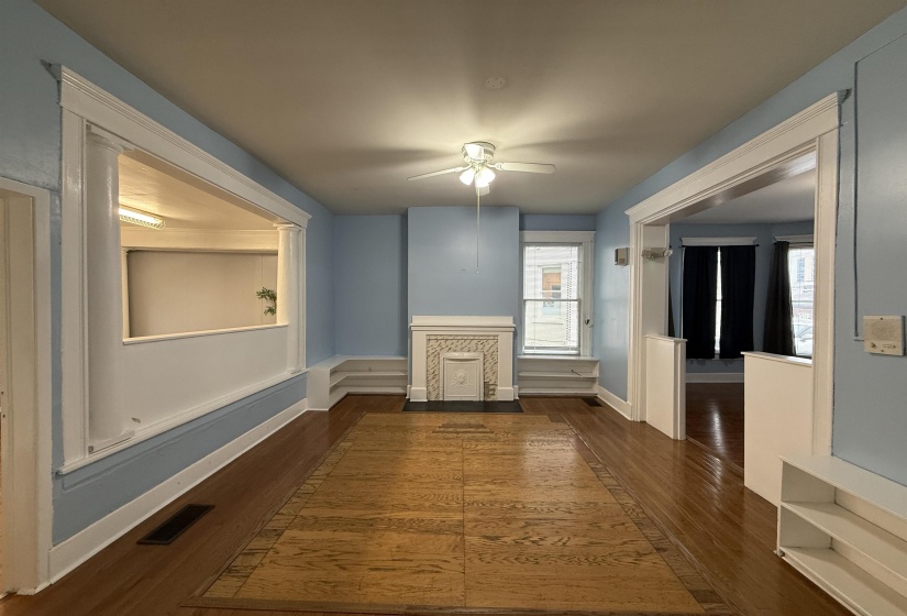 Spacious room featuring wood flooring with a decorative inlay, a ceiling fan light fixture, and a white decorative fireplace surround