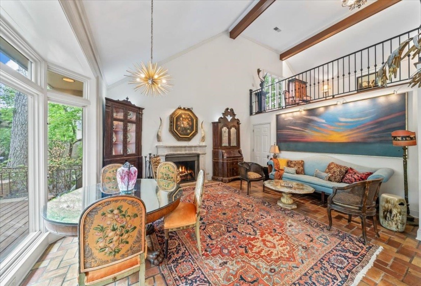 Keeping room with beamed ceiling, a lit fireplace, a chandelier, brick floors, and high vaulted ceiling and floor to ceiling windows overlooking large patio and gardens.