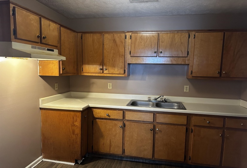 Kitchen with a textured ceiling, under cabinet range hood, light countertops, dark wood finished floors, and brown cabinetry