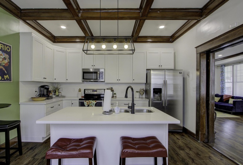 Kitchen featuring appliances with stainless steel finishes, hanging light fixtures, white cabinetry, a center island with sink, and beamed ceiling
